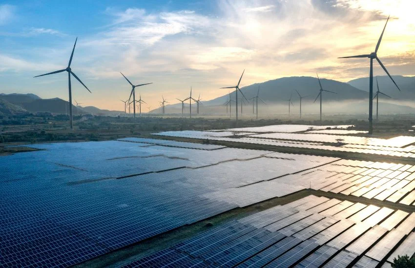 Paneles solares alineados en primer plano y varios molinos de viento girando en el horizonte bajo un cielo despejado al atardecer, representando el futuro de las energías renovables y la transición hacia fuentes de energía limpias y sostenibles.